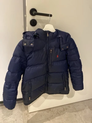 Mörkblå pufferjacka från Polo Ralph Lauren - Säljer jackan eftersom den har blivit för liten. Snygg mörkblå pufferjacka från Polo Ralph Lauren med röd logga på bröstet. Den ända defekten är att den har ett litet hål vid luvan.