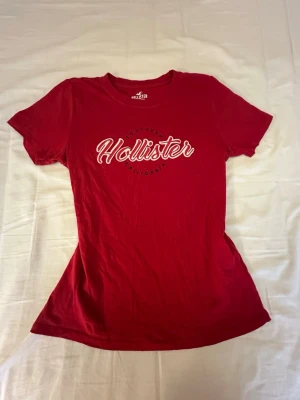Röd Hollister t-shirt med tryck - Snygg röd t-shirt från Hollister med vit logotyp och tryck på bröstet. Klassisk rund halsringning och korta ärmar. Tillverkad i mjuk bomull för skön känsla och passform. Perfekt för dig som gillar en enkel men stilren look.