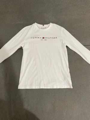 Vit långärmad topp Tommy Hilfiger - Säljer en vit långärmad topp från Tommy Hilfiger med tryckt logga och text 'EST. 1985' på bröstet. Klassisk rund halsringning och rak passform. Perfekt till jeans eller kjol för en clean och stilren look.