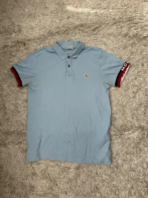 Ljusblå Moncler pikétröja - Säljer en ljusblå pikétröja från Moncler med klassisk krage och knappar framtill. Ärmarna har röda och svarta detaljer samt Moncler-logga på vänster bröst. Perfekt för en stilren och avslappnad look.