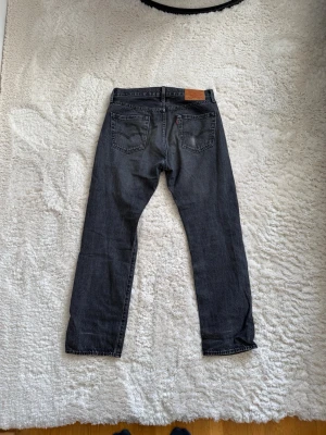 Svarta Levi's raka jeans 501 - Säljer ett par svarta raka jeans från Levi's med klassisk femficksmodell och silverfärgade nitar. Jeansen har en diskret tvättad look och är tillverkade i slitstark denim. Passar perfekt till en avslappnad streetstyle.