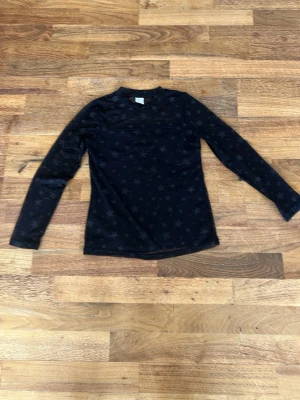 Svart mesh topp med stjärnmönster - Snygg svart mesh topp med diskret stjärnmönster. Toppen har lång ärm och är tillverkad i ett tunt, transparent syntetmaterial som ger en cool och trendig look. Perfekt att styla lager på lager eller bära som den är.