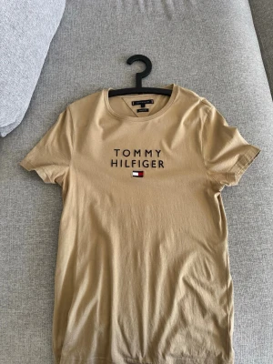 Beige t-shirt från Tommy Hilfiger - Snygg beige t-shirt från Tommy Hilfiger med klassisk logga och broderad flagga på bröstet. T-shirten har rund halsringning och korta ärmar. Perfekt basplagg med stilren design.