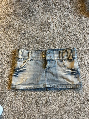 Blå jeanskjol med broderade fickor - Säljer en kort blå jeanskjol med slitningar och dubbla knappar framtill. Kjolen har broderade bakfickor och bälteshällor, perfekt för en avslappnad och trendig look. Klassisk femficksmodell i denim. Storlek S/xs