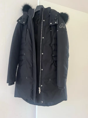 Svart parkas från Moose Knuckles - Snygg svart parkas från Moose Knuckles med avtagbar huva och svart fuskpäls. Jackan har dragkedja och knappar framtill, två fickor och logga på ärmen. Perfekt för kalla dagar med sin långa modell och vadderade design. (Kvitto finns)