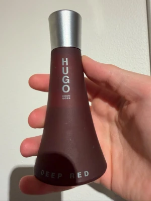 Hugo Boss Deep Red parfym 75ml flaska - Hugo Boss Deep Red är en parfym i en mörkröd, matt glasflaska med silverfärgat lock. Flaskan har en elegant, avsmalnande form och texten 'HUGO' och 'DEEP RED' i vitt. En klassisk doft som sticker ut med sin snygga design. Cirka 45-50ml kvar