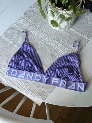 Lila bralette från Frank Dandy - Säljer en lila bralette från Frank Dandy med justerbara axelband och bred resår med logga. Triangelformade kupor med ränder och flamingomönster. Mjuk och bekväm i mesh utan bygel, perfekt för en avslappnad stil.