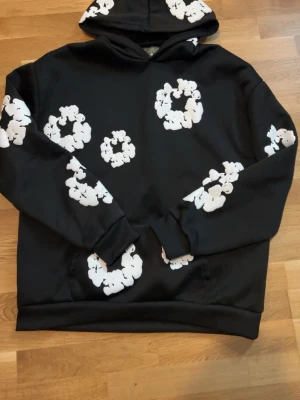 Denim & Tears - Säljer en svart hoodie med stora vita blomliknande tryck över hela tröjan, inklusive huvan och ärmarna. Hoodien har klassisk passform, huva och magficka. Perfekt för dig som gillar streetwear och vill sticka ut med ett unikt mönster.