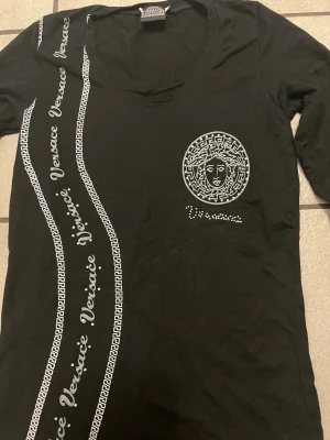 Svart långärmad topp från Versace storlek S - Snygg svart långärmad topp från Versace Jeans Couture med rund halsringning och diskret logga i nacken. Tillverkad i mjuk bomull som känns skön mot huden. Perfekt för dig som gillar stilrena och exklusiva plagg.