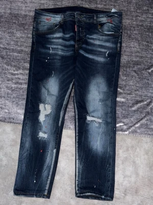 Mörkblå slitna jeans med detaljer - Säljer ett par mörkblå jeans med slitningar och färgstänk för en cool, avslappnad look. Jeansen har raka ben, klassisk femficksmodell och tydliga slitningar på både lår och knän. Materialet är denim i bomull. Perfekta för dig som gillar streetstyle.