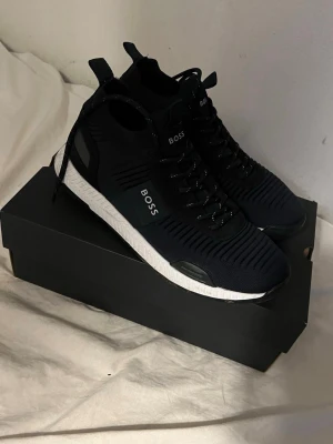  sneakers från BOSS - Stilrena svarta sneakers från BOSS med vit sula och stickad ovandel. Skorna har svarta skosnören med vita detaljer och BOSS-logga på sidan. Modern design med ribbade partier och bekväm passform. Perfekta för dig som gillar en clean och sportig look.