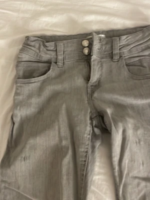 Gråa jeans med dubbla knappar - Säljer ett par gråa jeans med raka ben och dubbla knappar i midjan. Klassisk femficksmodell med bälteshällor och snygg tvättad look. Materialet känns mjukt och stretchigt, perfekt för en avslappnad stil.