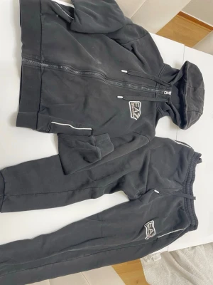 Svart EA7 loungewear set - Mysigt svart loungewear set från EA7 Emporio Armani med matchande hoodie och byxor. Setet har dragkedja på tröjan, huva och vita detaljer samt EA7-logga på både överdel och byxa. Perfekt för chill dagar hemma.