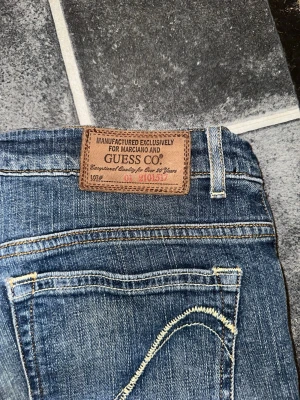 Blå jeans från Guess med kontrastsömmar - Säljer ett par klassiska blå jeans från Guess med snygga kontrastsömmar och femficksmodell. Jeansen har en normal midja och raka ben. Tillverkade i slitstark denim med läderpatch bak i midjan. Perfekta för en avslappnad och trendig look.