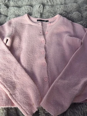 Ljusrosa kofta från 157 - Mysig ljusrosa kofta från 157 med rund halsringning och knappar framtill. Koftan har en mjuk, lite fluffig textur och långa ärmar. Perfekt för lager på lager och enkel att matcha med olika outfits.