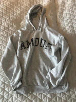 Grå hoodie med tryck 'AMOUR' - Snygg ljusgrå hoodie med svart text 'AMOUR' på bröstet och en stjärna på ryggen. Hoodien har huva och är från Gina tricot. Pris går att diskutera☺️