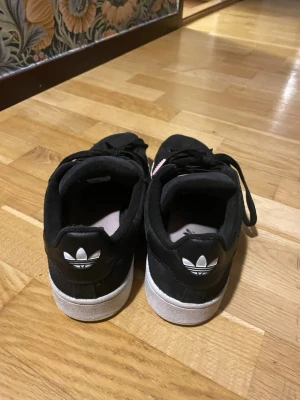 Svarta/rosa Adidas sneakers med vit sula - Snygga svarta sneakers från Adidas med klassisk vit sula och den ikoniska treklöver-loggan bak på hälen. Skorna har lågt skaft och svart ovandel i textil eller mocka. Perfekta för en avslappnad streetstyle-look.