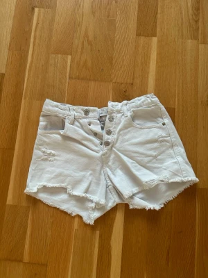 Vita denimshorts med knappar - Säljer ett par vita denimshorts med hög midja och råa, fransiga kanter. Shortsen har flera silverfärgade knappar framtill, slitna detaljer och klassiska fickor både fram och bak. Perfekta för sommaren och riktigt snygga till en avslappnad stil.