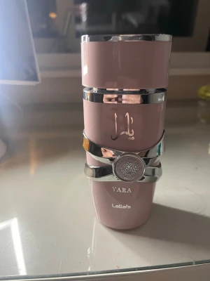 Lattafa Yara parfym rosa flaska - Lattafa Yara parfym i en snygg rosa flaska med silverdetaljer och dekorativt emblem runt mitten. Flaskan har en modern, cylindrisk form och arabiska bokstäver på framsidan. Perfekt för dig som gillar exklusiva och trendiga dofter. 95% kvar
