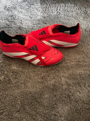 Adidas Predator röda fotbollsskor - Säljer ett par röda Adidas Predator fotbollsskor med vita och svarta detaljer. Skorna har klassisk låg profil, snörning och slitstark syntetisk ovandel. Perfekta för dig som vill ha grym kontroll på planen och snygg design.
