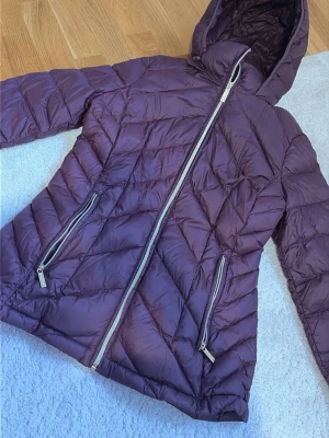 Lila pufferjacka med huva - Snygg lila pufferjacka med huva och sneddraget blixtlås framtill. Jackan har två dragkedjeförsedda fickor och quiltat mönster. Insidan är mörkare och materialet är lätt och vadderat, perfekt för kalla dagar.