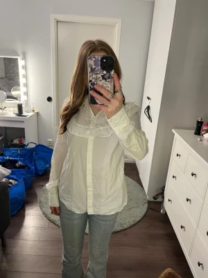 Vit skjorta med volangdetalj - Säljer en vit skjorta med volangdetalj över bröstet och klassisk krage. Skjortan har långa ärmar och knäppning framtill. Perfekt för dig som gillar en stilren men ändå söt look.