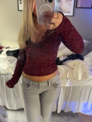Vinröd spetsblus med v-ringning - Vinröd långärmad blus i spets med v-ringad hals och croppad passform. Toppens spetsdetaljer ger en romantisk vibe och den sitter tight mot kroppen. Perfekt för dig som vill ha en trendig och feminin look.