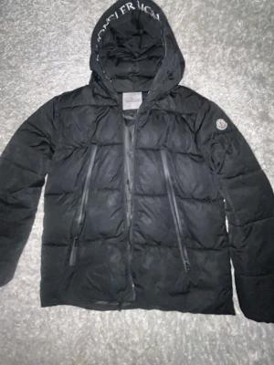 Svart dunjacka från Moncler - Säljer en svart dunjacka från Moncler med huva och två stora dragkedjefickor framtill. Jackan har Moncler-logga på ärmen och tryckt text på huvan. Insidan har ett serietidningsmönster och märkestaggar. Säljer den eftersom att jag använde den bara under vintern