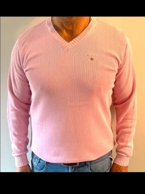 Ljusrosa stickad tröja från Gant - Snygg ljusrosa stickad tröja från Gant med V-ringning och diskret logga broderad på bröstet. Tröjan har långa ärmar och ribbade muddar vid ärmslut och nederkant. Perfekt för dig som gillar stilrena och klassiska plagg.