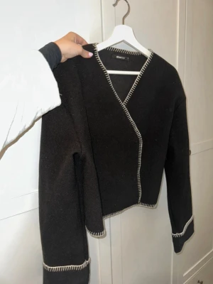 Svart omlottkofta med kontrastsömmar - Svart omlottkofta med vita kontrastsömmar längs kanter och ärmslut. Koftan har v-ringning, lång ärm och en loose passform. Materialet ser ut att vara ull och modellen är enkel men trendig, perfekt för lager på lager.