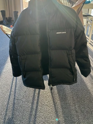 Svart pufferjacka från Arigato - Säljer en svart pufferjacka från Arigato med tryck 'ARIGATO ©2014' på bröstet. Jackan har hög krage, dragkedja framtill och två sidofickor med dragkedja. Klassisk boxig passform och tjock vaddering som håller dig varm under kalla dagar.