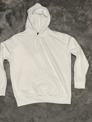 Vit basic hoodie med ficka - En stilren vit hoodie med huva och dragsko, samt en stor känguruficka framtill. Tröjan har ribbade muddar vid ärmslut och nederkant. Perfekt för en avslappnad och clean look. Materialet känns mjukt och bekvämt mot huden.