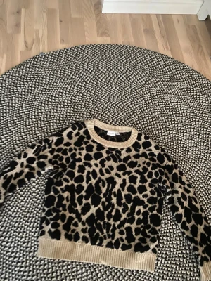 Leopardmönstrad stickad tröja - Säljer en snygg stickad tröja med leopardmönster i beige och svart. Tröjan har rund halsringning, ribbade muddar vid ärmslut och nederkant samt lång ärm. Perfekt för dig som vill sticka ut med ett djurmönster i garderoben.