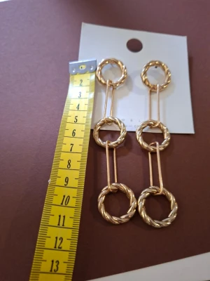 Guldiga statement örhängen i ringar - Säljer ett par långa statement örhängen i guldfärgad metall. Örhängena består av tre tvinnade ringar som sitter ihop med raka stavar, vilket ger en cool och trendig look. Perfekta för dig som vill sticka ut med accessoarerna.