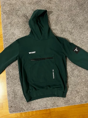 Mörkgrön hoodie från WOXO 720 - Snygg mörkgrön hoodie från WOXO 720 med stor magficka och dragkedja framtill. Tryck på bröstet och ärmen samt huva. Tillverkad i mjukt bomullsmaterial, perfekt för en avslappnad streetwear-look.