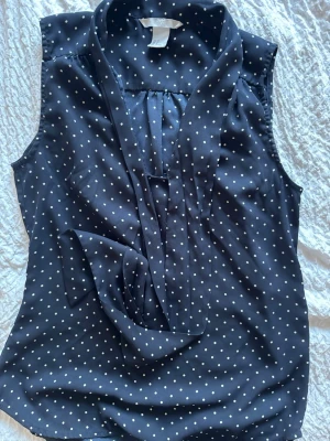 Mörkblå prickig blus från H&M - Snygg mörkblå blus från H&M med vita små prickar. Blusen är ärmlös och har en dekorativ knytning framtill samt en mjuk krage. Tillverkad i ett lätt och luftigt material som känns skönt mot huden. Perfekt för en trendig och avslappnad look.