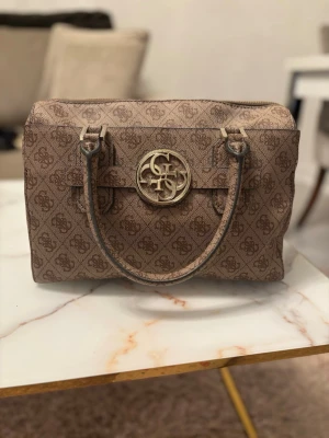 Beige handväska från Guess - Snygg beige handväska från Guess med klassiskt monogrammönster och guldfärgad logga framtill. Väskan har två stadiga handtag och en boxig form som ger plats för allt du behöver. Tillverkad i konstläder med detaljer i metall.