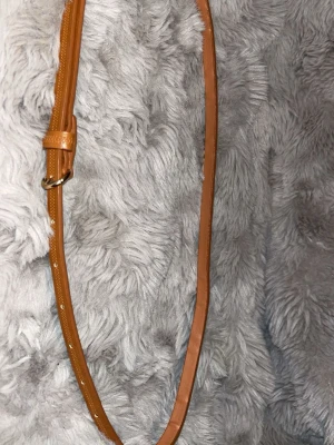 Smalt brunt bälte i skinn - Snyggt och smalt bälte i brunt skinn med guldfärgat spänne. Perfekt för att ge din outfit en stilren touch. Bältet har små hål för justering och en enkel design som passar till många olika looks.