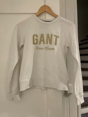 Vit sweatshirt från GANT - Vit sweatshirt från GANT med guldfärgad text 'GANT New Haven' på bröstet. Tröjan har rund halsringning, långa ärmar och ribbade muddar vid ärmslut och nederkant. Perfekt för en avslappnad och stilren look.