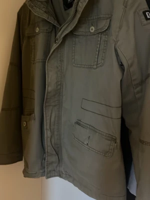 Olivgrön jacka från Brandit - Snygg olivgrön jacka från Brandit med flera fickor framtill och knappdetaljer. Jackan har hög krage och dragkedja samt tryckknappar. Perfekt för dig som gillar en avslappnad och funktionell stil.