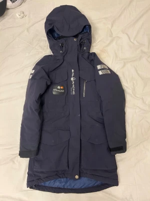 Mörkblå Sail Racing parkas Gore-Tex - Mörkblå parkas från Sail Racing med GORE-TEX-material, stor huva och flera praktiska fickor. Jackan har logotyp på bröstet och ärmen samt patch med texten 'Arctica Expedition'. Lång modell med dragkedja och kardborre vid ärmslut. Perfekt för kalla dagar.