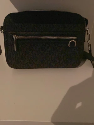 Svart axelväska från Michael Kors - Snygg svart axelväska från Michael Kors med silverfärgade detaljer och dragkedja framtill. Väskan har ett diskret MK-mönster över hela ytan och justerbar axelrem. Perfekt för dig som vill ha en stilren och praktisk väska till vardagen.