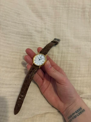 Klassisk guldklocka med brunt äkta läderband - Stilren klocka med vitt ur, guldfärgad boett och brunt äkta läderarmband med krokodilpräglad yta. Urtavlan har romerska siffror och datumvisning. Perfekt accessoar för dig som gillar tidlös design och klassiska detaljer.