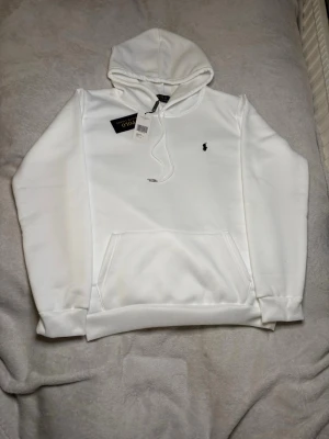 Vit hoodie från Polo Ralph Lauren - Snygg vit hoodie från Polo Ralph Lauren med klassisk svart broderad logga på bröstet. Hoodien har huva med dragsko, magficka och ribbade muddar vid ärmslut och nederkant. Tillverkad i mjukt bomullsmaterial för skön känsla.