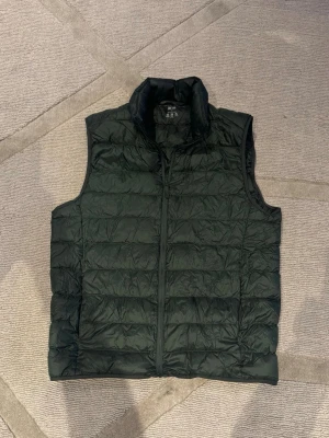 Uniqlo väst - Snygg mörkgrön dunväst med ståkrage och quiltad design. Västen har dragkedja framtill och är lätt vadderad för extra värme. Perfekt att slänga över en hoodie eller tröja när det är vår ute. Enkel och stilren look som passar till det mesta.