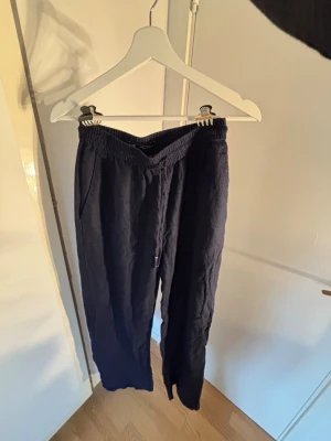 Mörkblå linnebyxor med dragsko - Snygga mörkblå linnebyxor med avslappnad passform och elastisk midja med dragsko. Byxorna har sidfickor och en bakficka, perfekta för en chill och stilren look. Tillverkade i luftigt linnematerial som känns skönt mot huden.