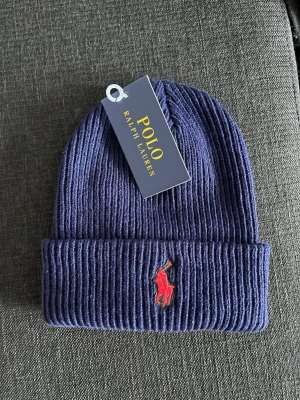 Marinblå ribbad mössa Polo Ralph Lauren - Snygg marinblå ribbstickad mössa från Polo Ralph Lauren med klassisk röd broderad logga framtill. Perfekt accessoar för kyliga dagar och enkel att matcha med olika outfits.