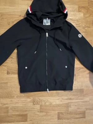 Svart vindjacka från Moncler - Snygg svart vindjacka från Moncler med huva och dragkedja framtill. Jackan har två fickor med tryckknappar, dragsko i huvan och Moncler-logga på ärmen. Huvan har en röd och vit rand som detalj. Perfekt för en sportig och stilren look.