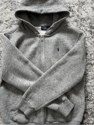 Grå hoodie från Polo Ralph Lauren - Snygg grå hoodie från Polo Ralph Lauren med dragkedja och blå broderad logga på bröstet. Hoodien har mjukt, vitt fleecefoder på insidan och ribbade muddar vid ärmar och nederkant. 
