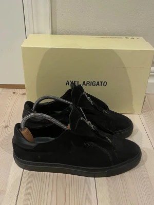 Svarta sneakers från Axel Arigato - Stilrena svarta sneakers från Axel Arigato i mocka med diskret logga på sidan. Skorna har en klassisk låg siluett och svart sula. Insidan är fodrad med läder för extra komfort. Perfekta för dig som gillar minimalistisk och modern design. Säljer då dem är för småa